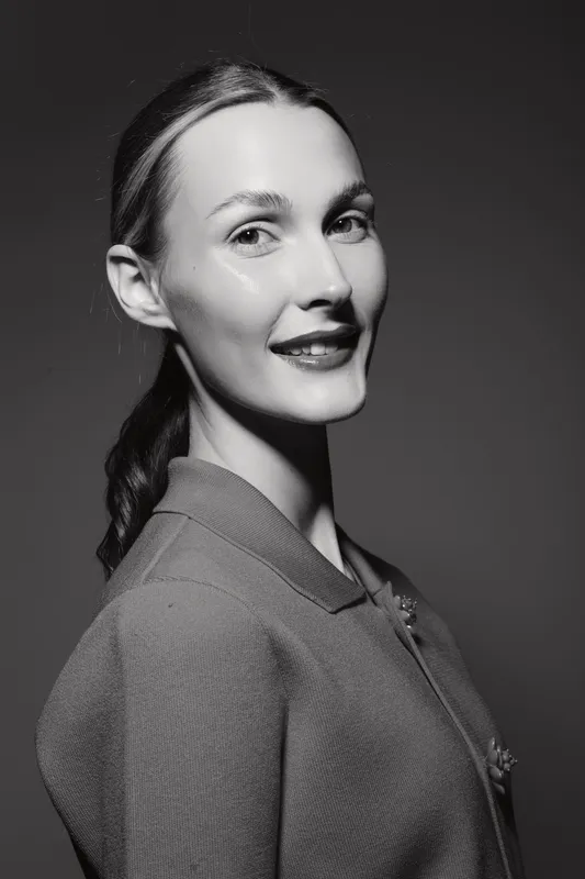 Portrait of a smiling Caucasian woman with pale skin and slicked back hair in a dark textured jacket. Studio lighting highlights her facial features on black background.