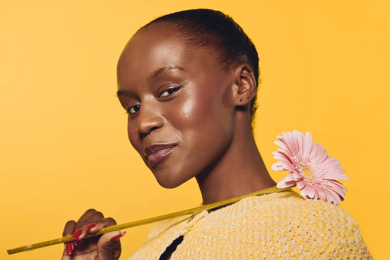 Portrait of a confident dark skinned African woman holding a pink flower on her shoulder. She wears a yellow sweater against a bright yellow background. Concept of beauty and natural elegance.