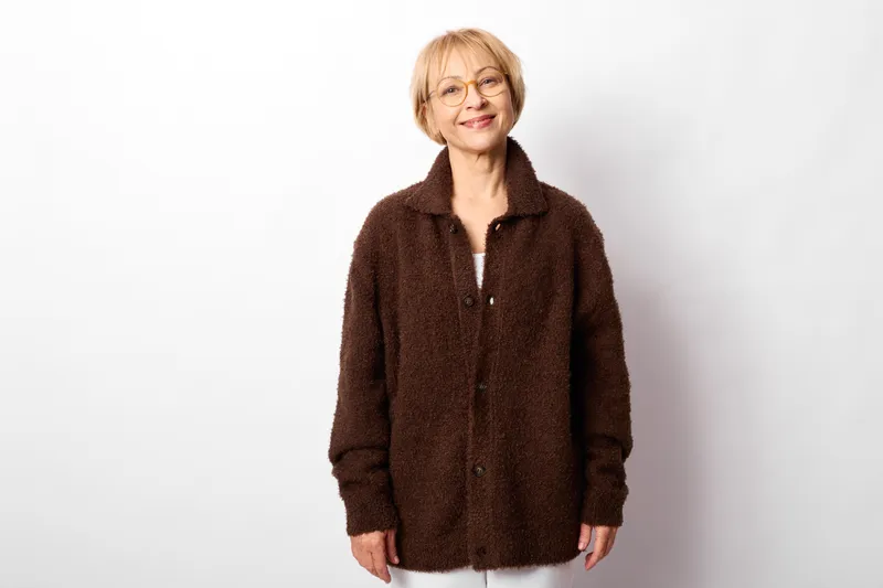 Elderly woman smiling with glasses in studio portrait, wearing brown jacket on white background, relaxed pose, cozy everyday fashion, soft lighting, calm mood