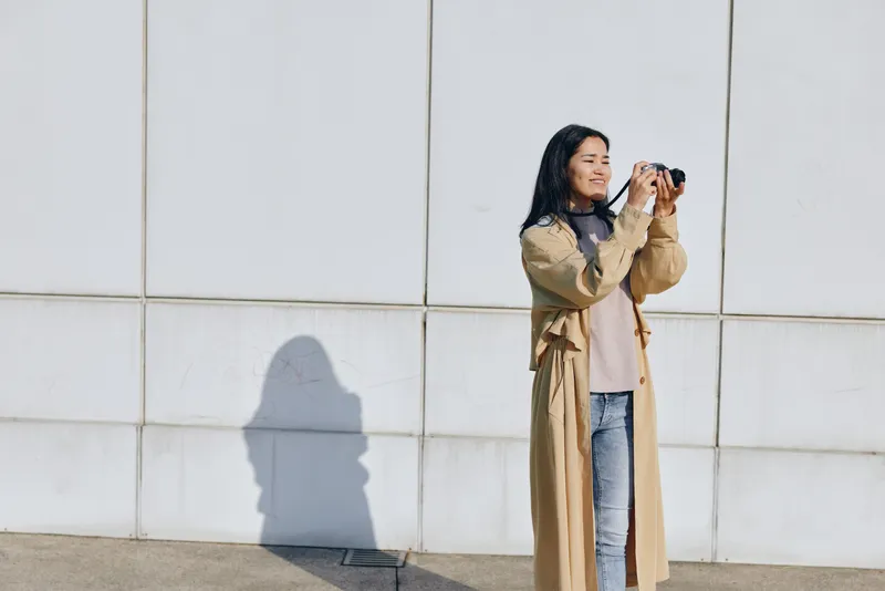 Woman photographer in a long beige coat captures a moment with camera outdoors against a white tiled wall, sunlight casts a long shadow, casual fashion style, candid urban composition.