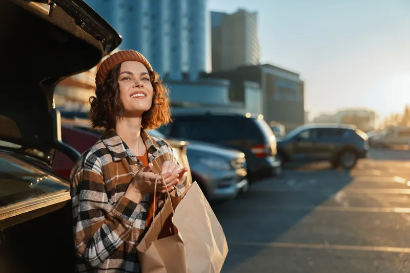 Woman shopping by car in urban parking, holding paper bags with a smile in golden hour sunlight, candid lifestyle portrait with authenticity, mindful living and emotional storytelling glow.