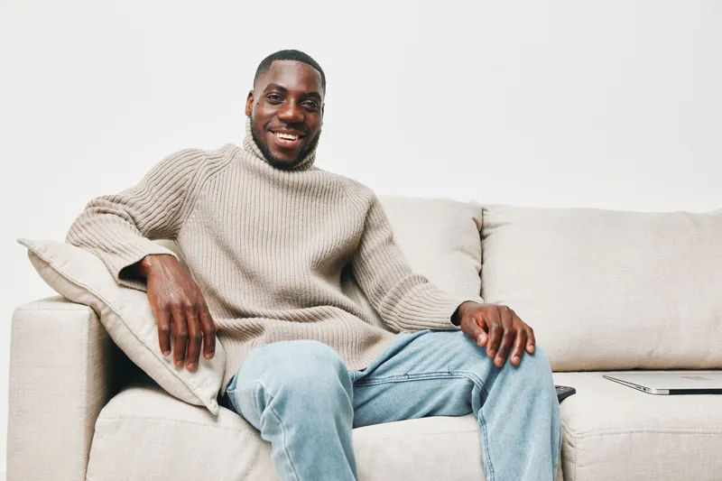 Smiling Black man in cozy sweater sitting on a sofa, expressing warmth and comfort, ideal for seasonal lifestyle themes and home decor images
