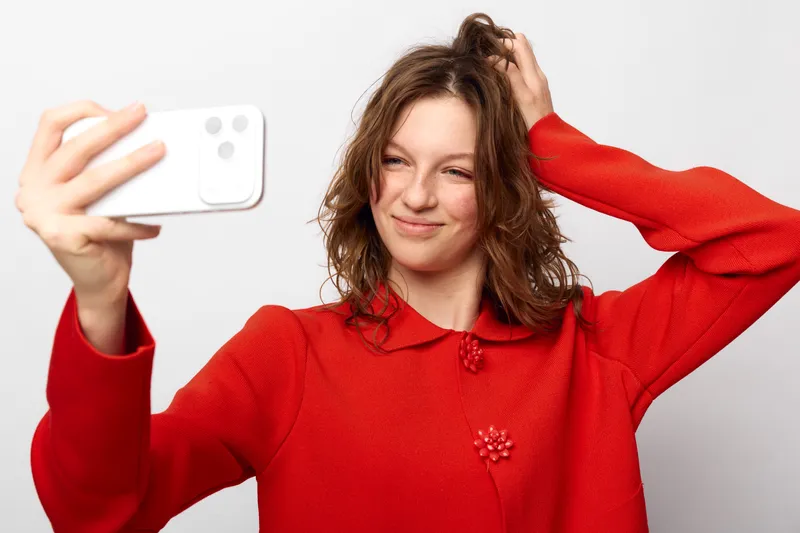 Young woman in red sweater takes a selfie with smartphone, smiling and touching hair. Studio portrait on white background. Concept of casual lifestyle, technology, and self-expression.