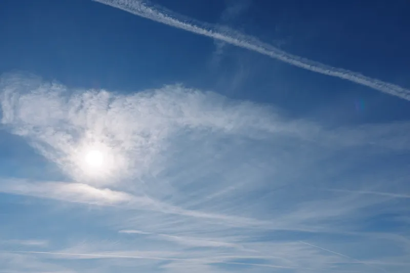 Clear blue sky with bright sun and wispy clouds, airplane contrails crossing overhead, creating a serene daytime scene suitable for nature, travel and outdoor lifestyle projects