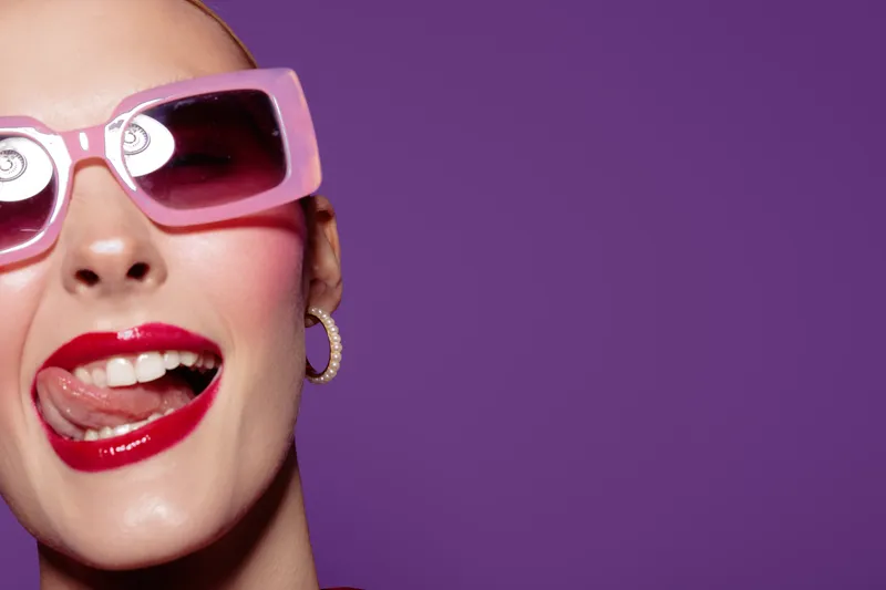 Close-up of a happy woman wearing pink sunglasses and red lipstick, smiling with tongue out. Bright purple background creates a fun and playful mood for fashion and beauty concept.