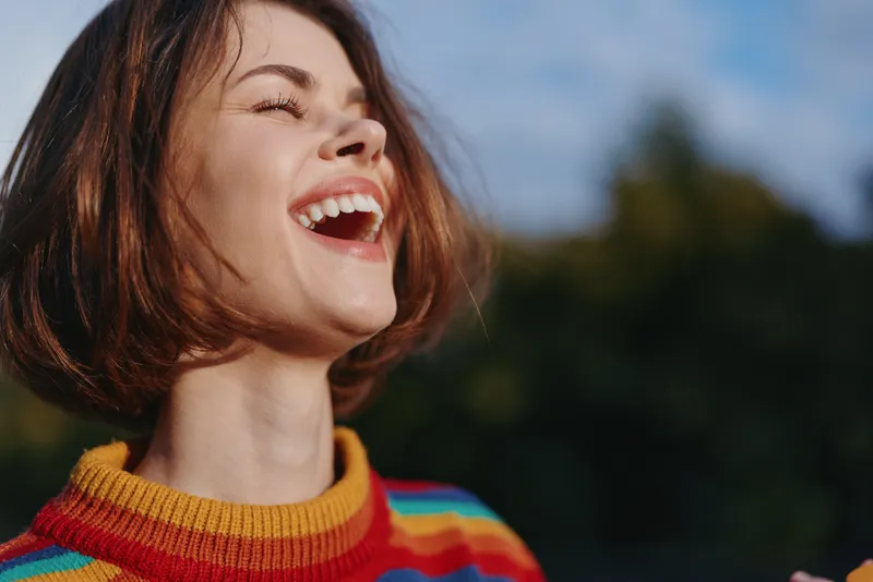 woman smile laughing striped sweater short hair outdoor sunlight portrait, mid 20s female laughing with open mouth, rainbow knit sweater, joyful lifestyle fashion portrait in warm golden hour nature