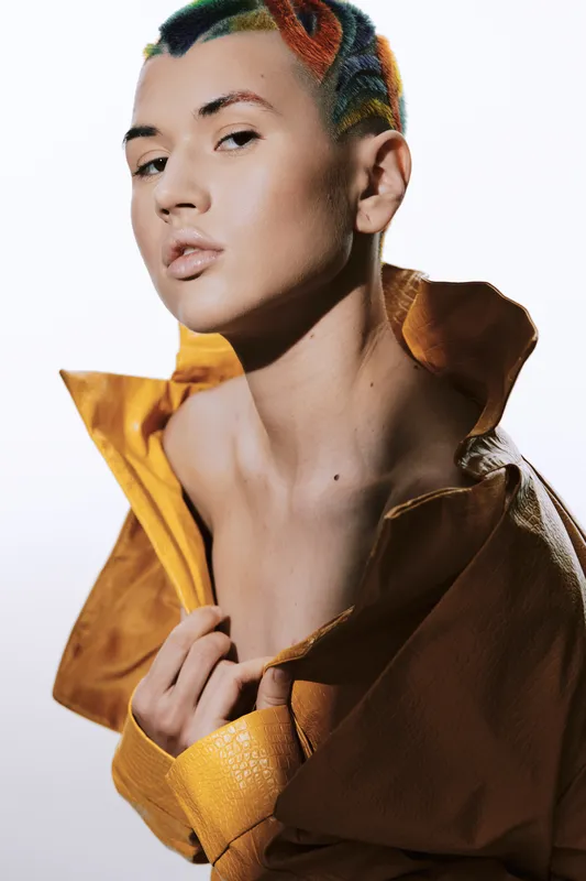 Stylish woman with short colorful hair and smooth skin poses wearing a bold yellow textured coat on a clean white background, showcasing modern fashion and beauty.