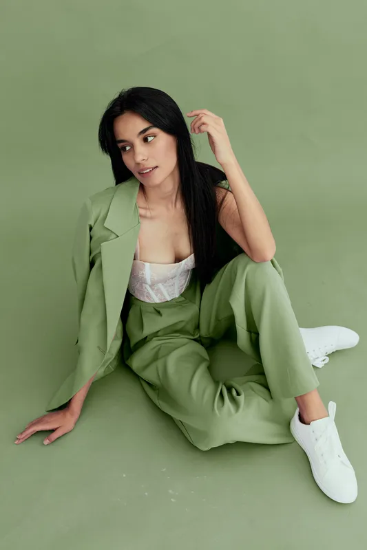 Stylish woman in green pantsuit sitting on floor against green background, wearing white sneakers and lace top, with long dark hair and relaxed posture, modern fashion look.