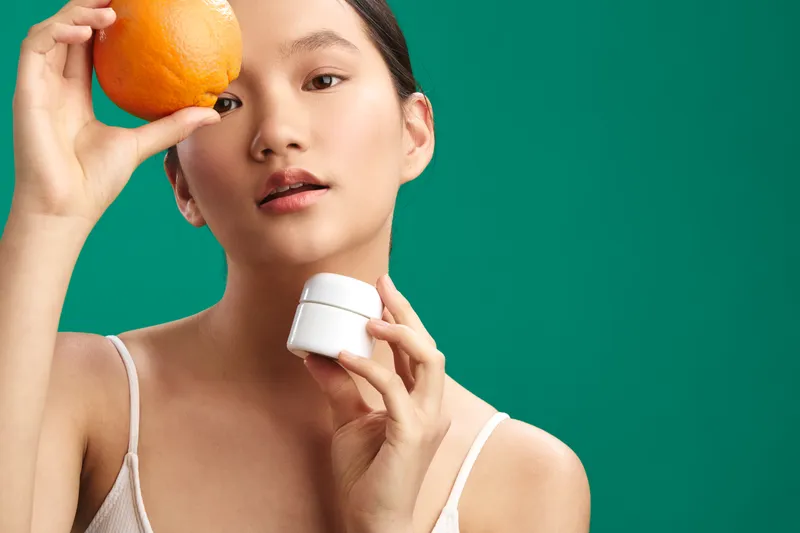 asian woman holds an orange near her face and a small cream jar in hand, standing before a vivid green backdrop to illustrate a fresh skincare moment and confident glow.