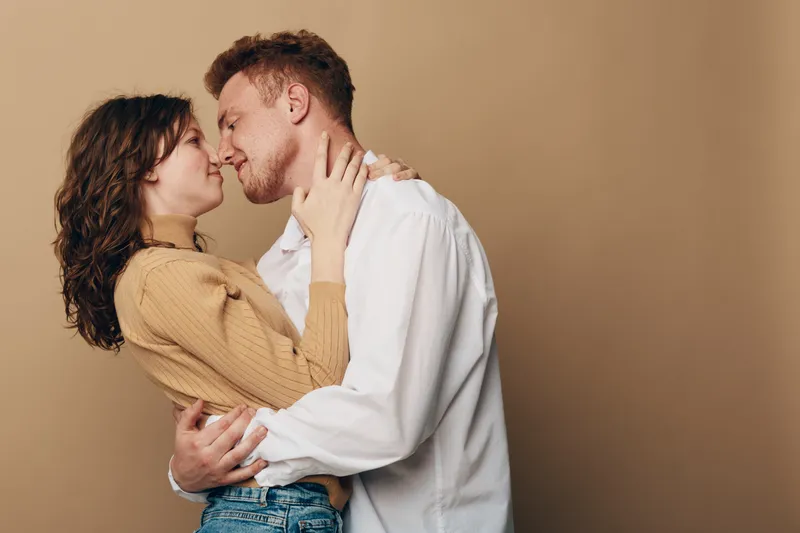 Young couple embraces closely, looking into each other's eyes with affection. Casual clothing and warm beige background create intimate romantic mood and connection.