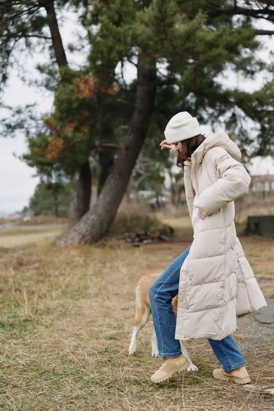 Person in a white puffer coat and beanie walking with a dog in a park, winter scene with trees and grass, casual outdoor activity today
