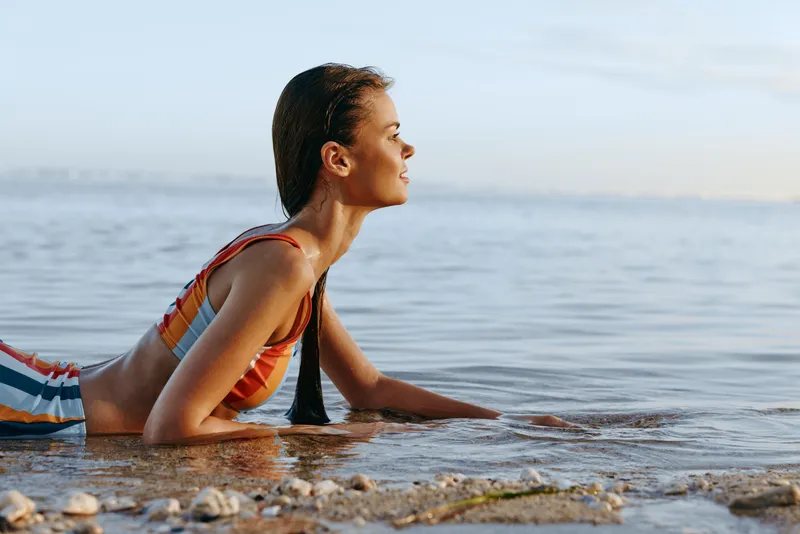 A young woman in a striped swimsuit lies on shallow water at the beach, propping her upper body with forearms as gentle waves lap the shore, conveying calm focus with a quiet shimmer
