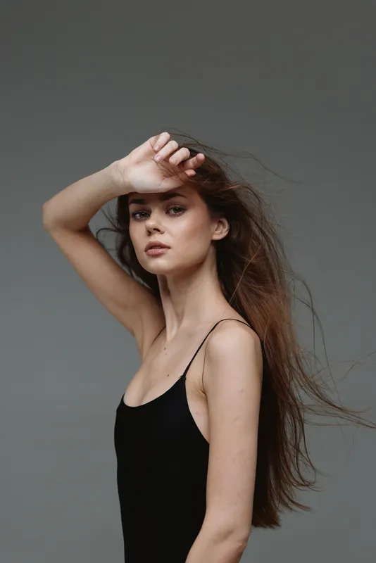 thoughtful woman posing with flowing hair in black dress against gray background, stylish beauty, serene mood