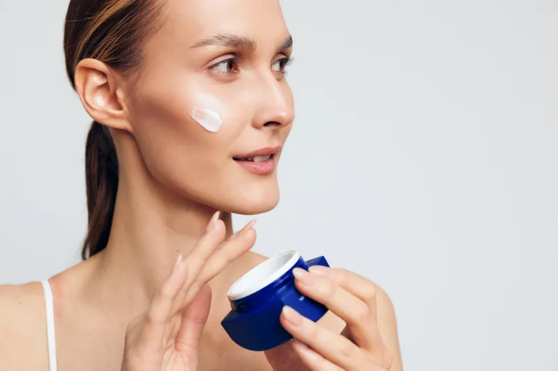 Young woman applying face cream with fingers, skincare routine, clear skin, natural beauty, soft lighting, white background, closeup portrait, healthy lifestyle, moisturizing product