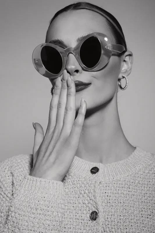 Elegant caucasian woman with light skin wearing large round sunglasses and knitted sweater covers mouth with hand. Studio portrait in black and white. Concept of fashion and style.