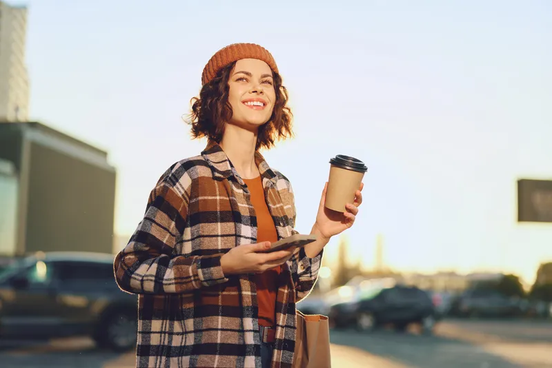 woman with coffee and smartphone on street smile in plaid jacket and beanie, sunlight creating golden hour glow, candid lifestyle portrait with authenticity and mindful living vibe.