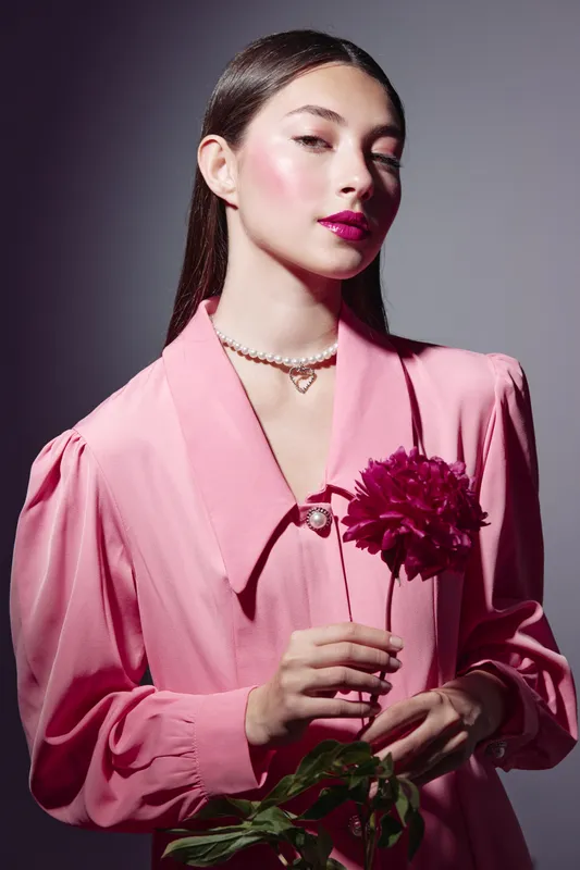 Young woman with sleek hair in a pink blouse holds a deep red flower with pearl necklace. Studio lighting highlights her glowing skin and bold makeup. Beauty fashion creative concept.