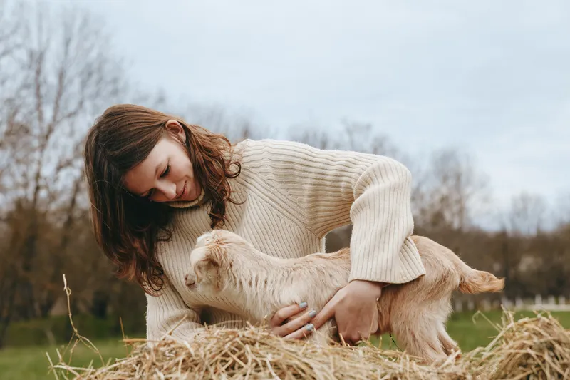 Calm woman in a chunky sweater gently strokes a young goat kid on a sunny farm, seated on hay, showcasing rural lifestyle, care, and harmony with nature.
