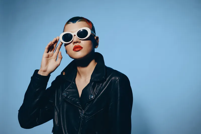 Stylish woman with shaved head wearing white round sunglasses and black leather jacket against blue background, showcasing strong fashion statement and bold attitude.