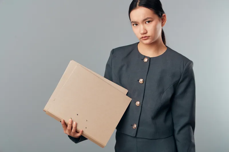 Young woman holding cardboard boxes in casual business outfit with serious expression on grey background. Indoor portrait of female carrying flat parcel for delivery or storage purpose.