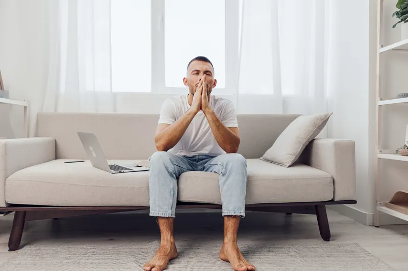 Concentrated man sitting on a cozy sofa with a laptop, showcasing the modern lifestyle of remote work in a bright, minimalistic room filled with natural light.