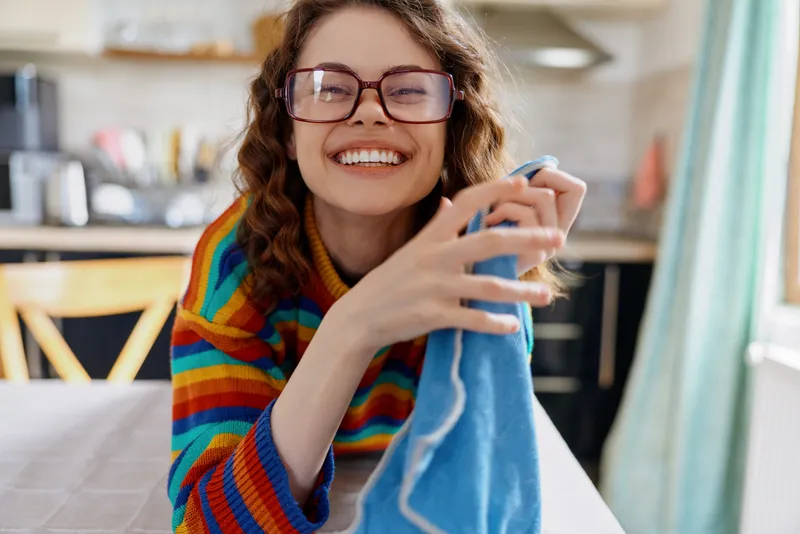 A cheerful woman with glasses in a striped sweater dries a towel in a bright kitchen, capturing a warm home moment and a simple cleaning routine.