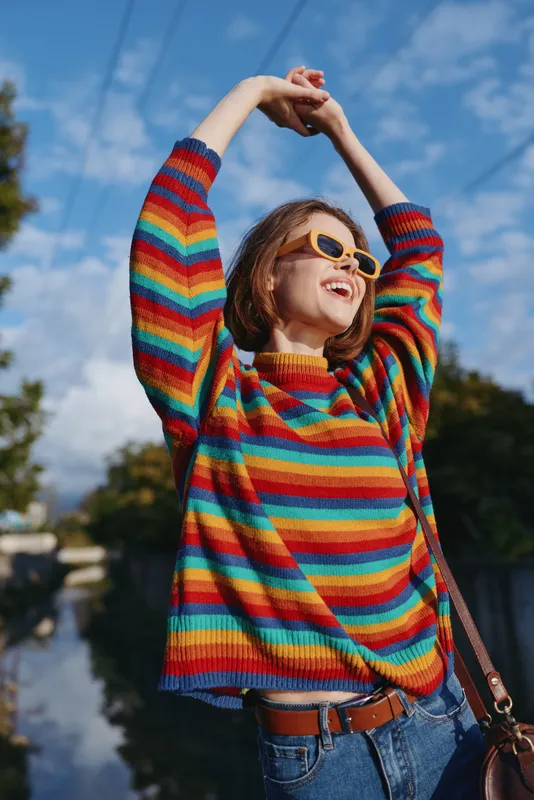Woman in colorful striped sweater and sunglasses with a bright smile, joyful outdoors portrait stretching arms up in casual urban style under sunny blue sky.