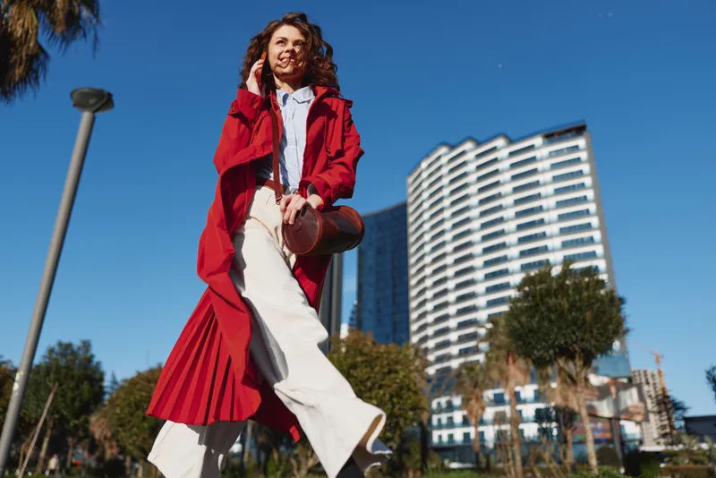 Young woman in a bright red coat walks confidently through a sunny city street, smiling as she strides past modern high rise buildings.