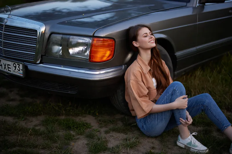 Young woman enjoying the outdoors by a vintage car, wearing casual denim and a loose shirt, exuding relaxation and tranquility in nature.