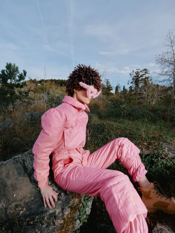 Pink jumpsuit worn by a model seated on a rock in an outdoor landscape, blending fashion and nature with a playful headband and casual pose for a vibrant editorial