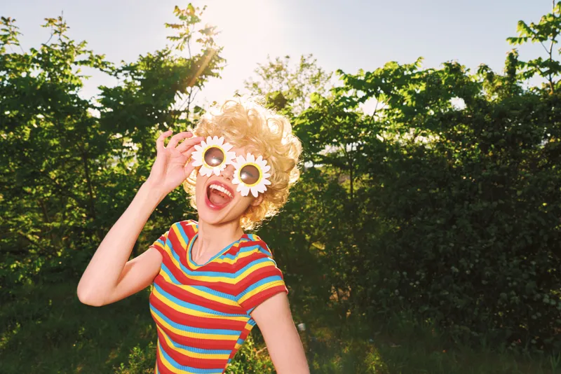 Playful woman outdoors wears a rainbow striped top and flower sunglasses, in a sunlit garden, capturing retro film color and a vibrant casual lifestyle moment Joyful,bright,energetic
