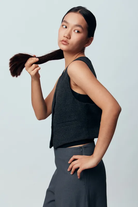 Young woman with long dark hair holding ponytail looking away in sleeveless black vest and gray pants on light background studio portrait.