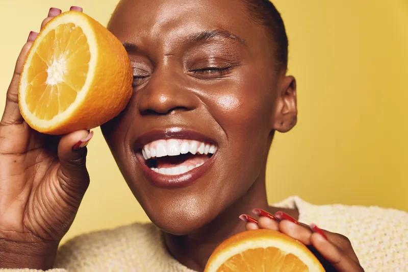 Happy African woman with dark skin holds fresh orange slices near face, smiling with closed eyes. Bright yellow background and natural makeup create fresh healthy concept.