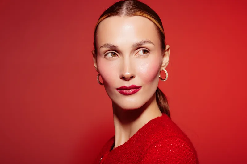 Portrait of a confident woman with slicked back hair and red lipstick wearing hoop earrings and a red sweater against a red background. Concept of fashion, beauty, and elegance.