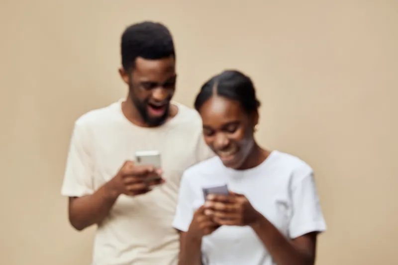 Happy young African couple engaged with smartphones, wearing casual white shirts, against a soft beige background, expressing joy and connection through technology