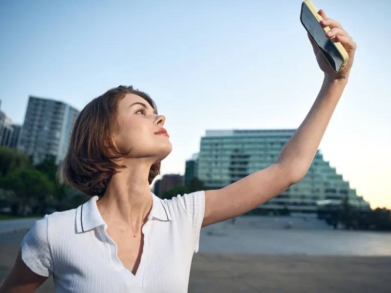 A confident woman holds a smartphone high, posing for a selfie on a sunny beach with modern buildings in the background, creating a cheerful, relaxed and upbeat outdoor mood.