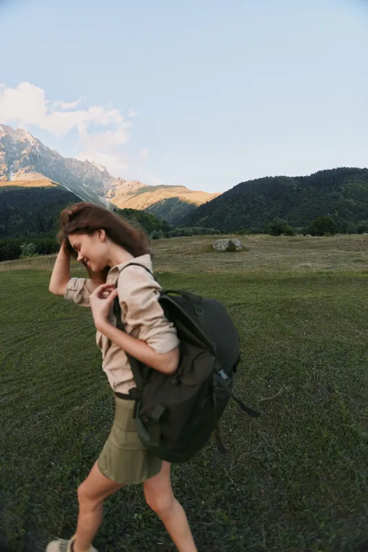 Young woman with a backpack walking through a meadow beneath mountains, showcasing outdoor lifestyle and wellness in a natural setting, with warm natural light and a candid sense of movement.