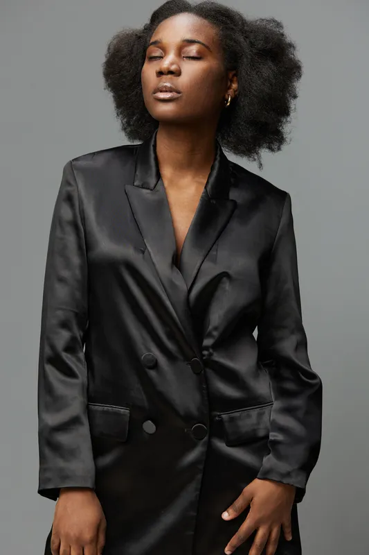 Elegant young woman with natural hair in a black satin blazer, showcasing confidence and poise against a neutral backdrop for fashion and beauty concepts