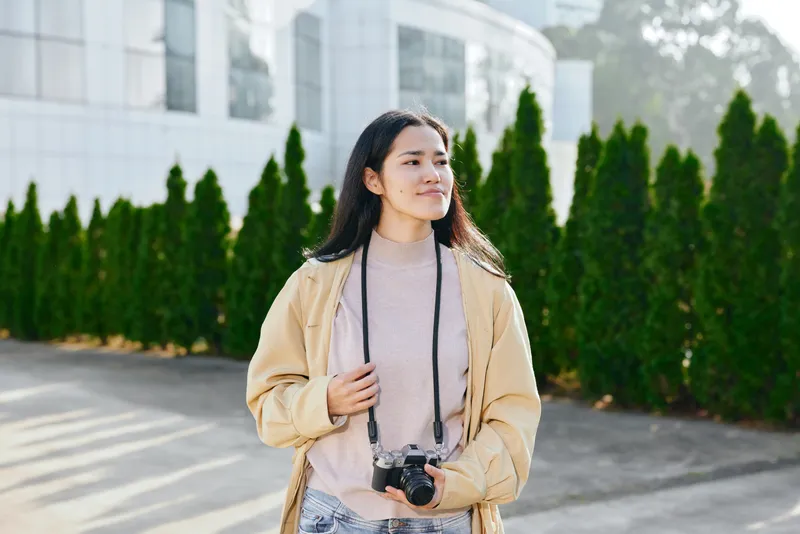 Camera in hand, a woman in a beige jacket stands outdoors under bright daylight, with green hedges and buildings in the background, ready for a casual photo shoot, and expresses calm focus for