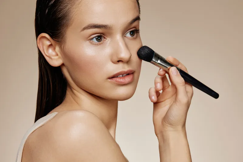 Elegant young woman applying makeup with a brush, showcasing natural beauty and a minimalist style against a soft beige background, ideal for beauty and skincare concepts