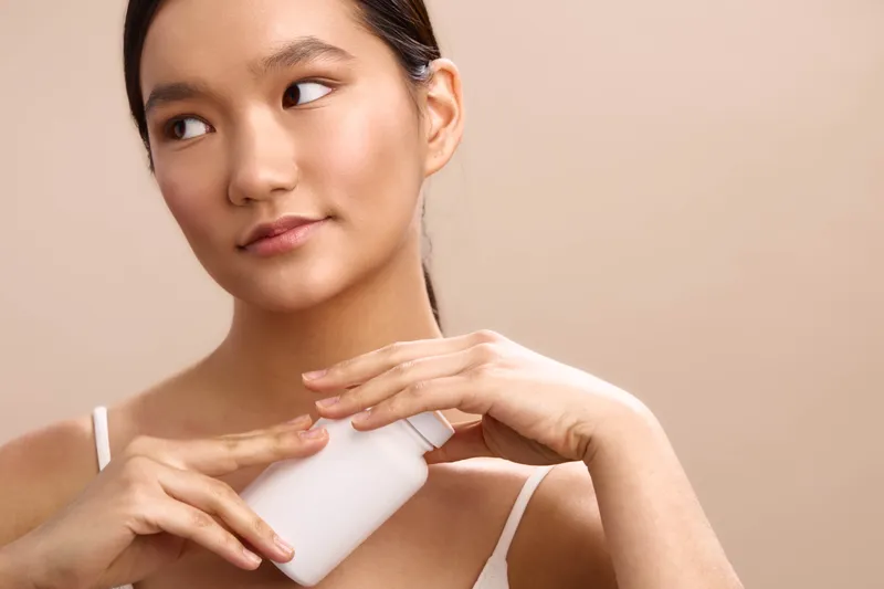 asian woman applies cream on her shoulder in a soft studio setting, showcasing a focused skincare moment. the calm expression and minimalist backdrop convey a peaceful personal care routine.