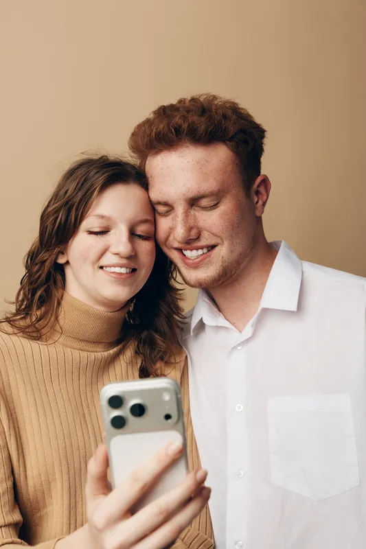 Young couple with curly hair smiling and looking at smartphone screen together. Casual clothing and warm beige background create cozy and happy atmosphere of shared moment.