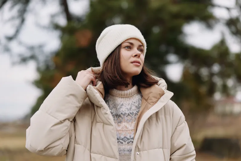 Woman wearing beige winter coat and knit sweater stands outdoors with a white beanie, soft natural light, calm expression, chilly vibe, rustic landscape and cozy fashion in a candid portrait