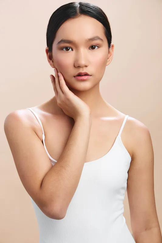 Asian woman portrait in a clean studio setting, wearing a white tank top. She gazes softly at the camera, conveying calm confidence and understated elegance.