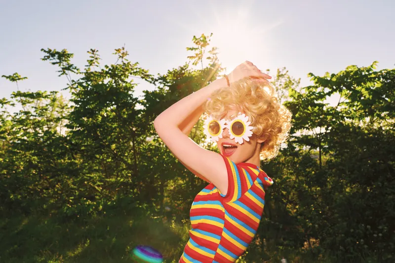 Playful young girl with curly blonde hair dances in a sunlit garden, wearing striped tee and oversized flower sunglasses, captured with retro film color for a vibrant summer vibe.
