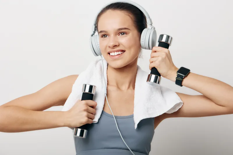 Woman fitness enthusiast wearing headphones, lifting dumbbells with a towel draped around the neck, energetic smile, studio backdrop, illustrating strength training and active lifestyle during a