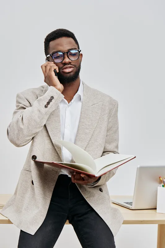 Professional young man with glasses wearing beige blazer and white shirt, holding a phone to his ear and reading a book, standing near a desk with a laptop in a bright office, business lifestyle conce