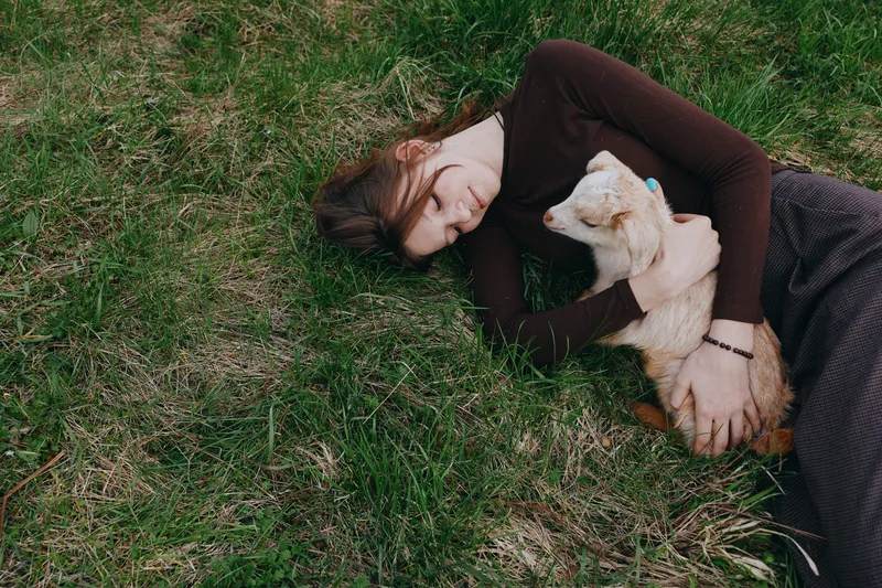 Relaxed outdoor moment, a woman lying on grass, embracing a gentle goat kid, sunlit, soft colors, peaceful mood, nature inspired lifestyle scene and textured grass beneath, soft breeze