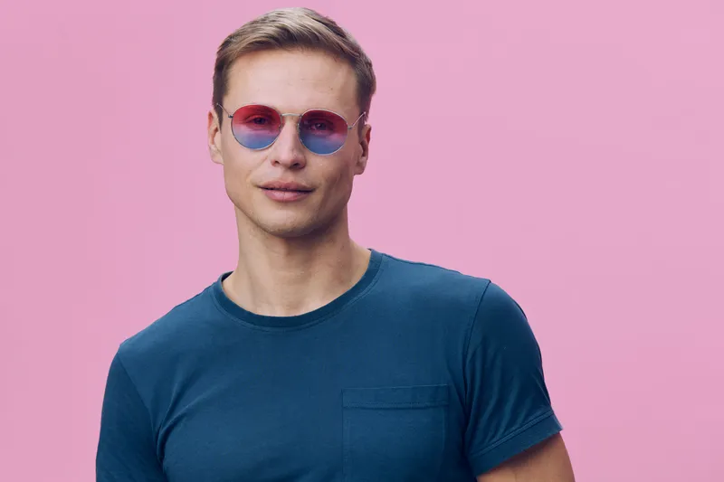 Man wearing pink sunglasses and blue t-shirt posing against a pink background, lifestyle image, casual style, young adult, cheerful expression, summer vibe.