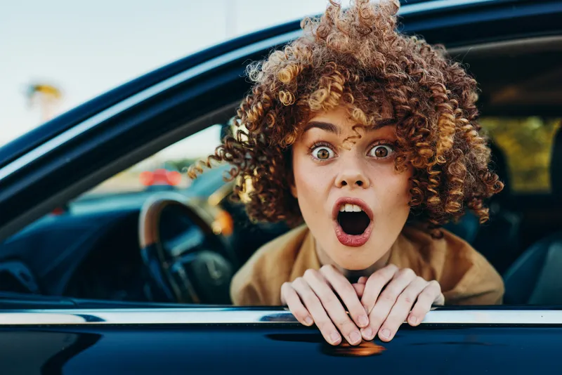 A woman with surprise and astonishment leans from a car window, creating a bold portrait. The lifestyle scene features retro film color, vivid light, and candid emotion.
